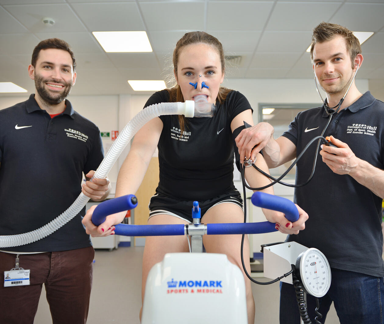 Woman on a stationary bike performing a fitness test with two male sport and exercise scientists from The University of Hull either side.