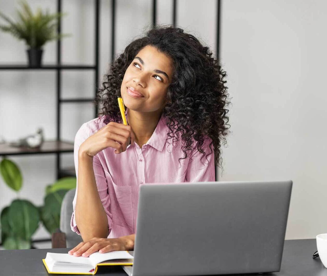 Young person sat at a desk with an open laptop, thinking and note taking.