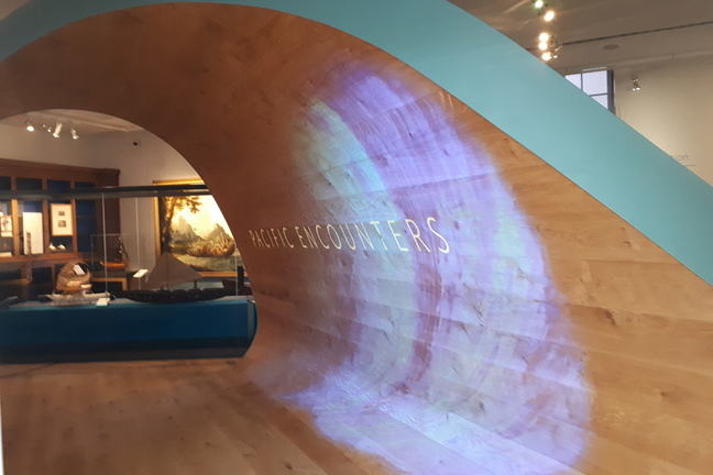 Entrance display to gallery with the words Pacific Encounters on a large wave-style wooden wall