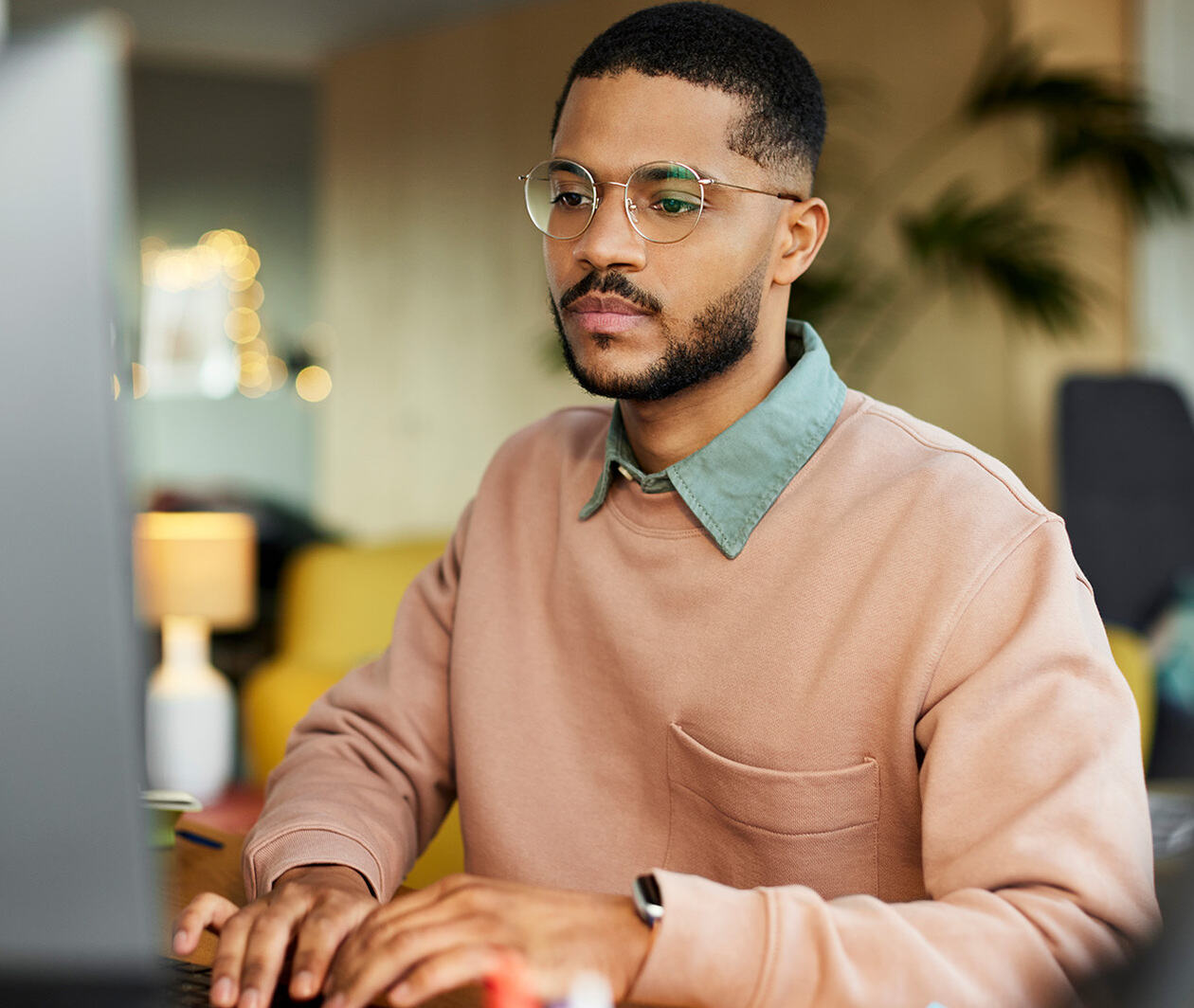 A man in a pink jumper and glasses is on a computer in an office setting.