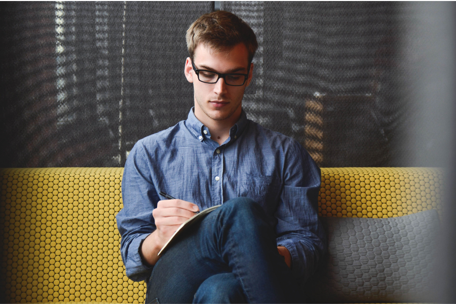 Young man with black glasses wearing light blue linen shirt and jeans seated on couch writing on notepad in lap. Couch is yellow with geometric pattern; cushion to man's left is grey with matching geometric pattern.