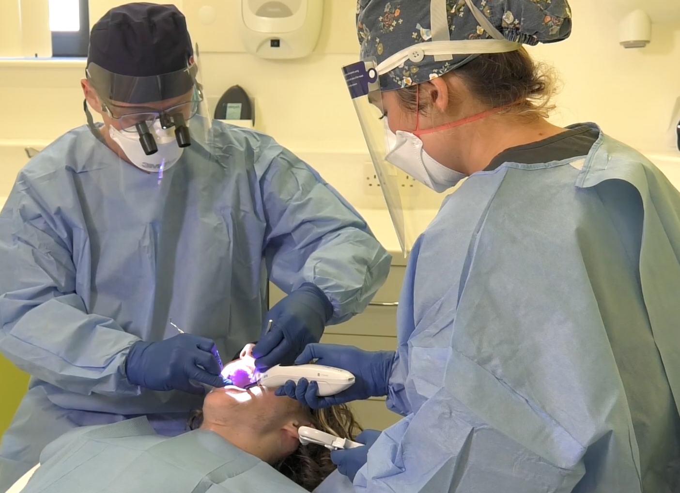 Photograph of two dentists wearing surgical hats, gowns, masks, glasses and face visors