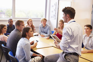 Students around a table listening to a man holding a tablet