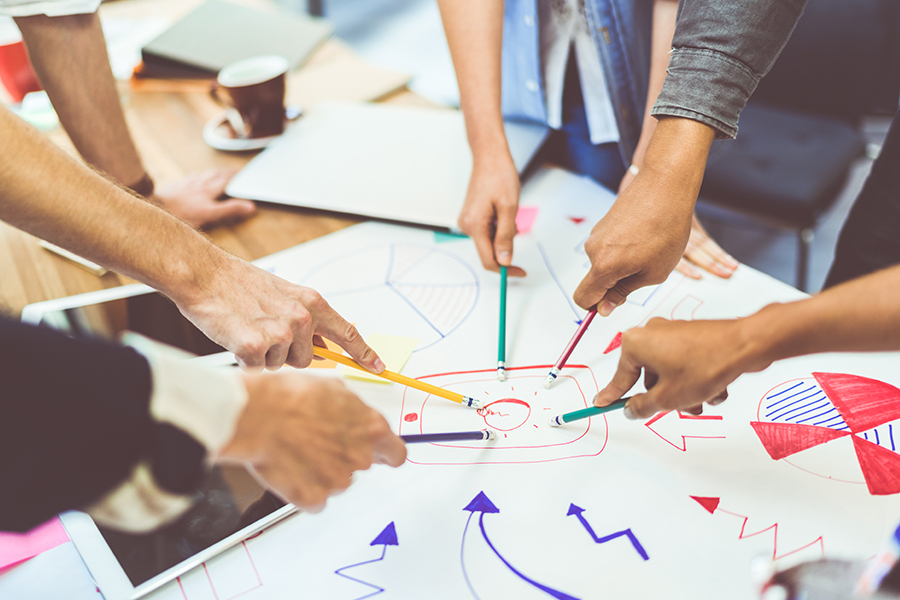 A group of people standing over a table, looking at a drawing of ideas and pointing toward the drawing with their pens.