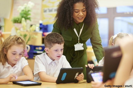 A female teacher leans over a desk where pupils are looking at tablet computers. The girl on the left holds a finger over the screen; the boy in the middle holds his tablet. Another girl is visible but out-of-focus on the right.