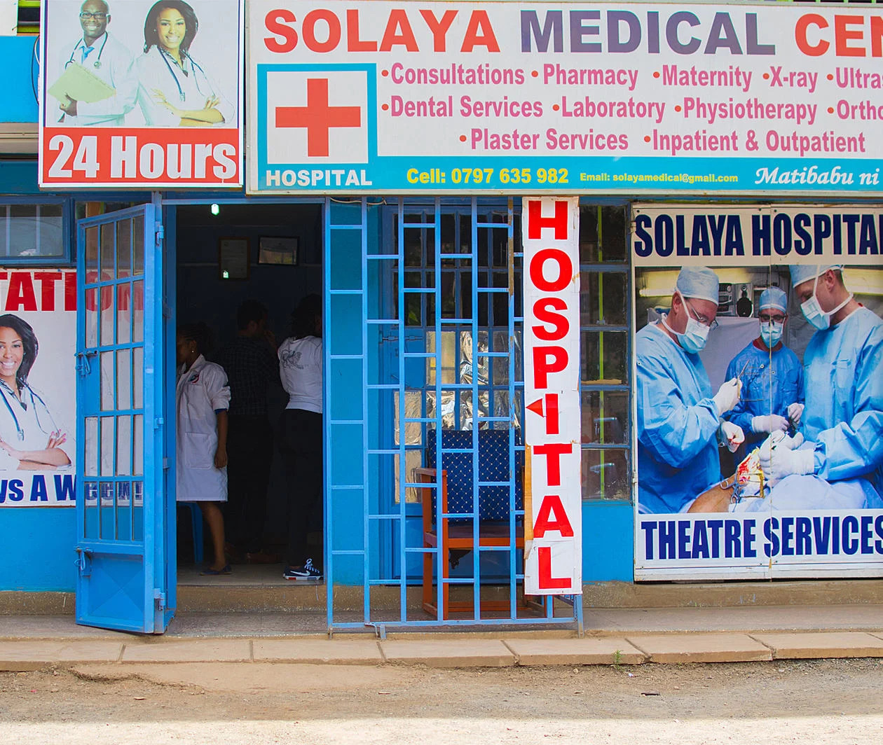 The entrance to a hospital called Solaya medical centre in Kenya. Building is blue with blue railings around and there is detail on the hospital signage about the services offered at the hospital which is a 24 hour 7 days a week hospital