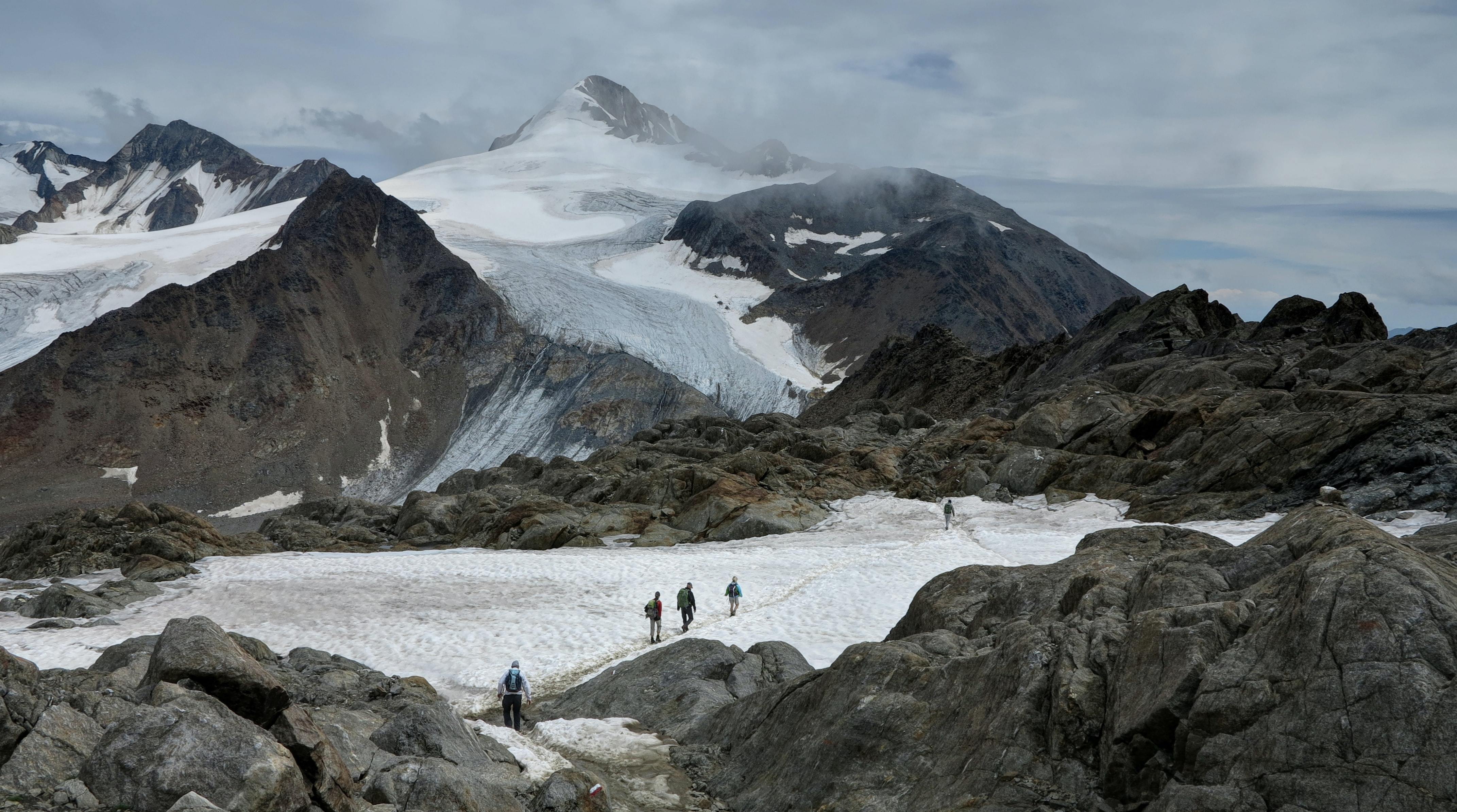 Image shows a group of people walking across a snow patch towards a glacier in the distance, surrounded by mountains.