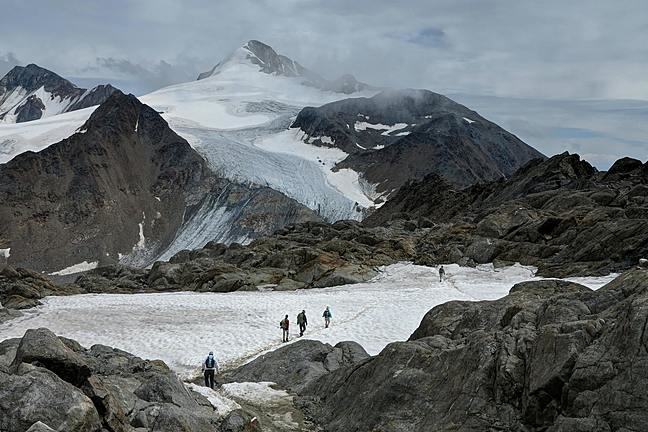 Image shows a group of people walking across a snow patch towards a glacier in the distance, surrounded by mountains.