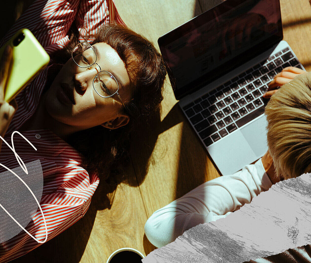 Two young people lying on the floor in the sunlight looking at a mobile phone and laptop.