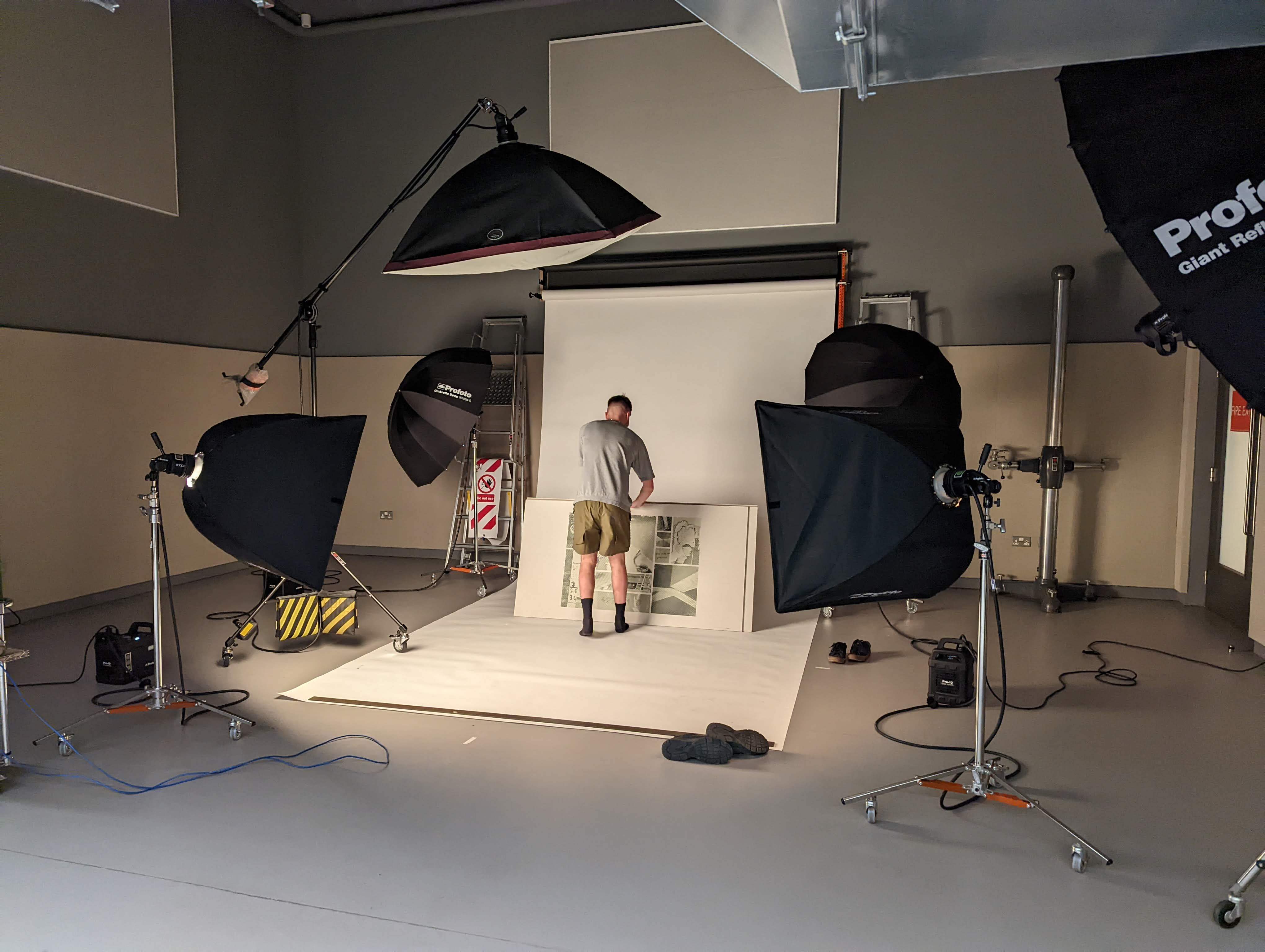 Student photographing their work in a room with white and grey walls. There are black lights set up and a white pull down backdrop.