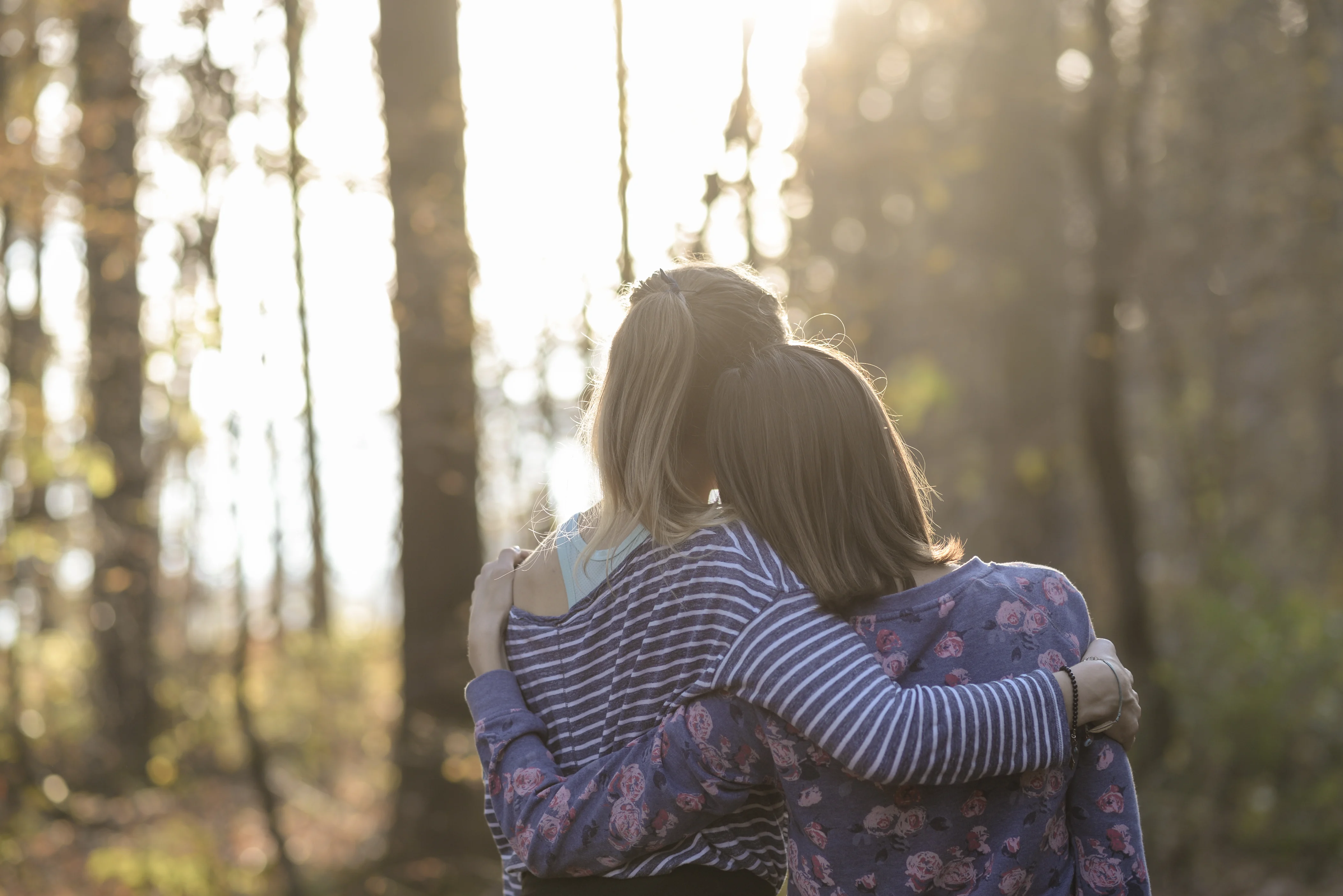 Two women walking in a forest with their arms around one another.