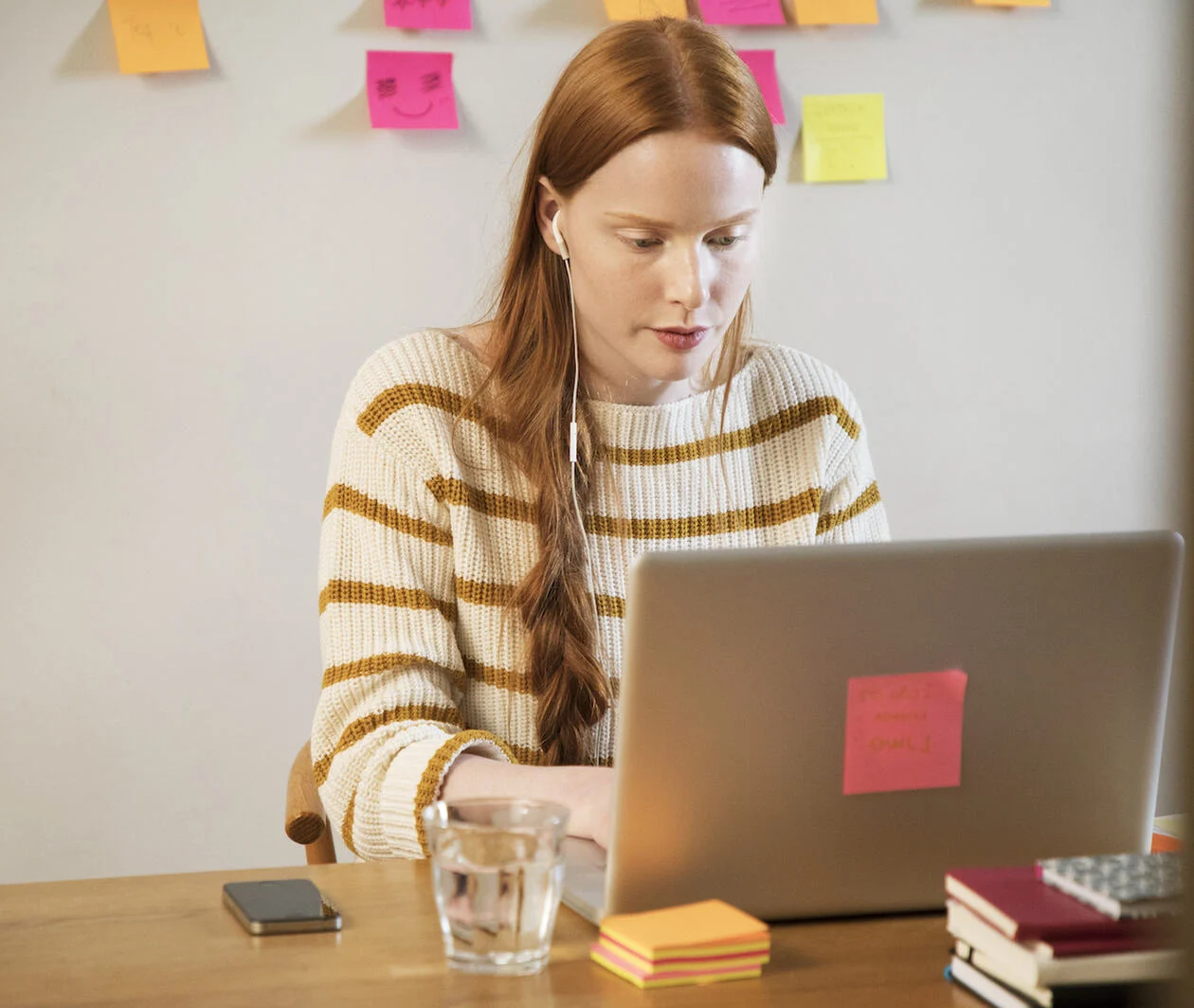 A young woman sat at a laptop, working on her research project, with post-its on the wall be hind her detailing her development