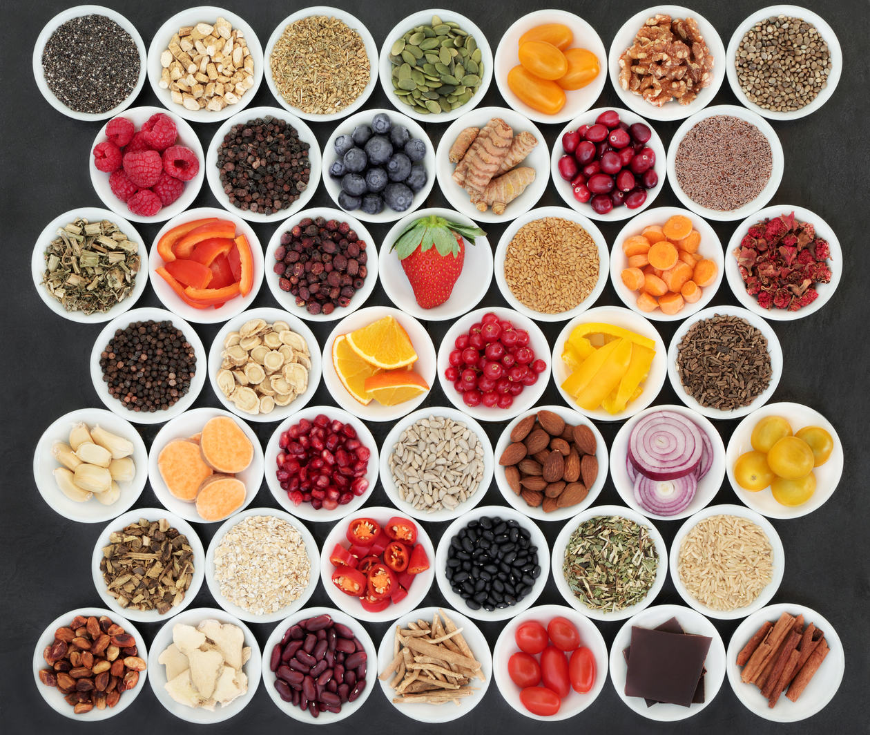 Rows of white bowls each containing different food to create a colourful pattern. Foods include raspberries, blueberries, tomatoes, cinnamon, red onion sliced and sliced peppers.