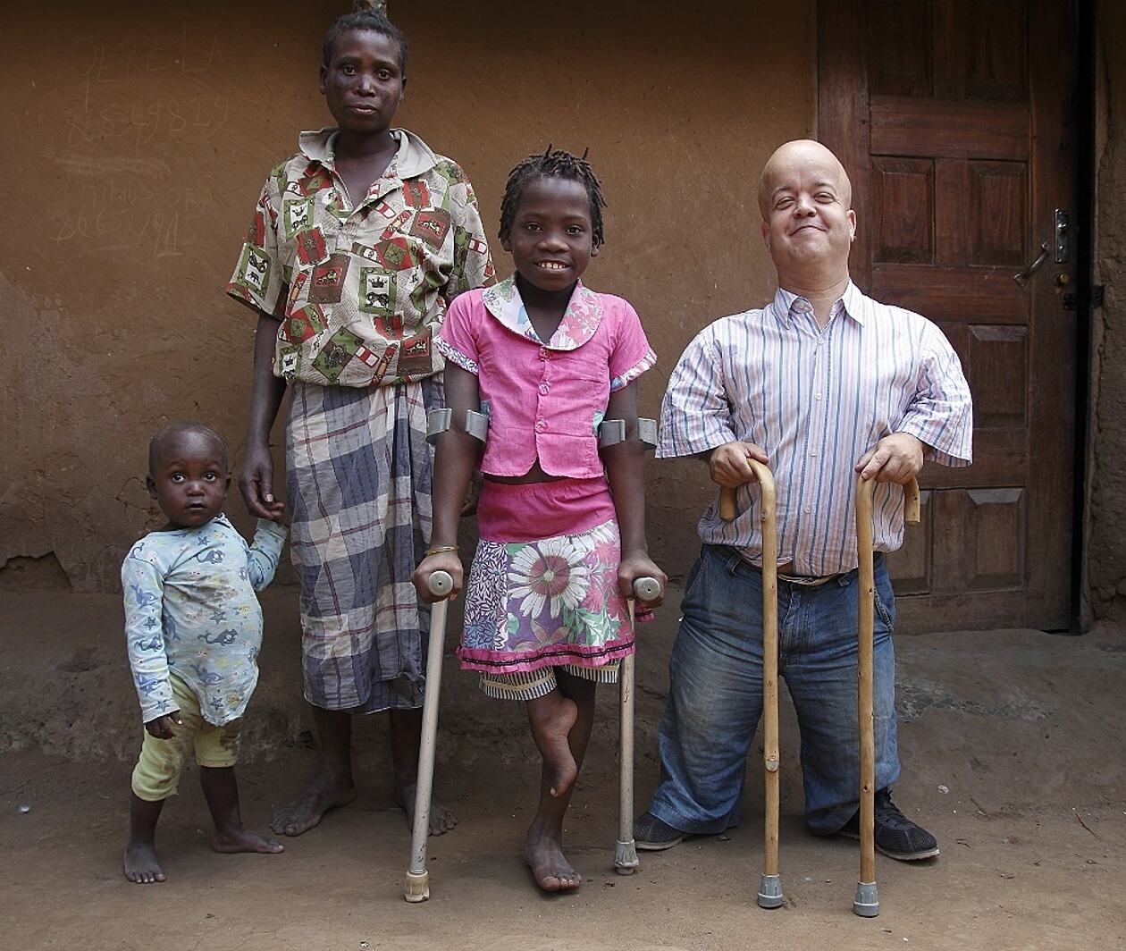 Image shows four people posing for camera, from left to right a child holding his mother’s hand, an older female child using crutches, and a man using walking sticks.