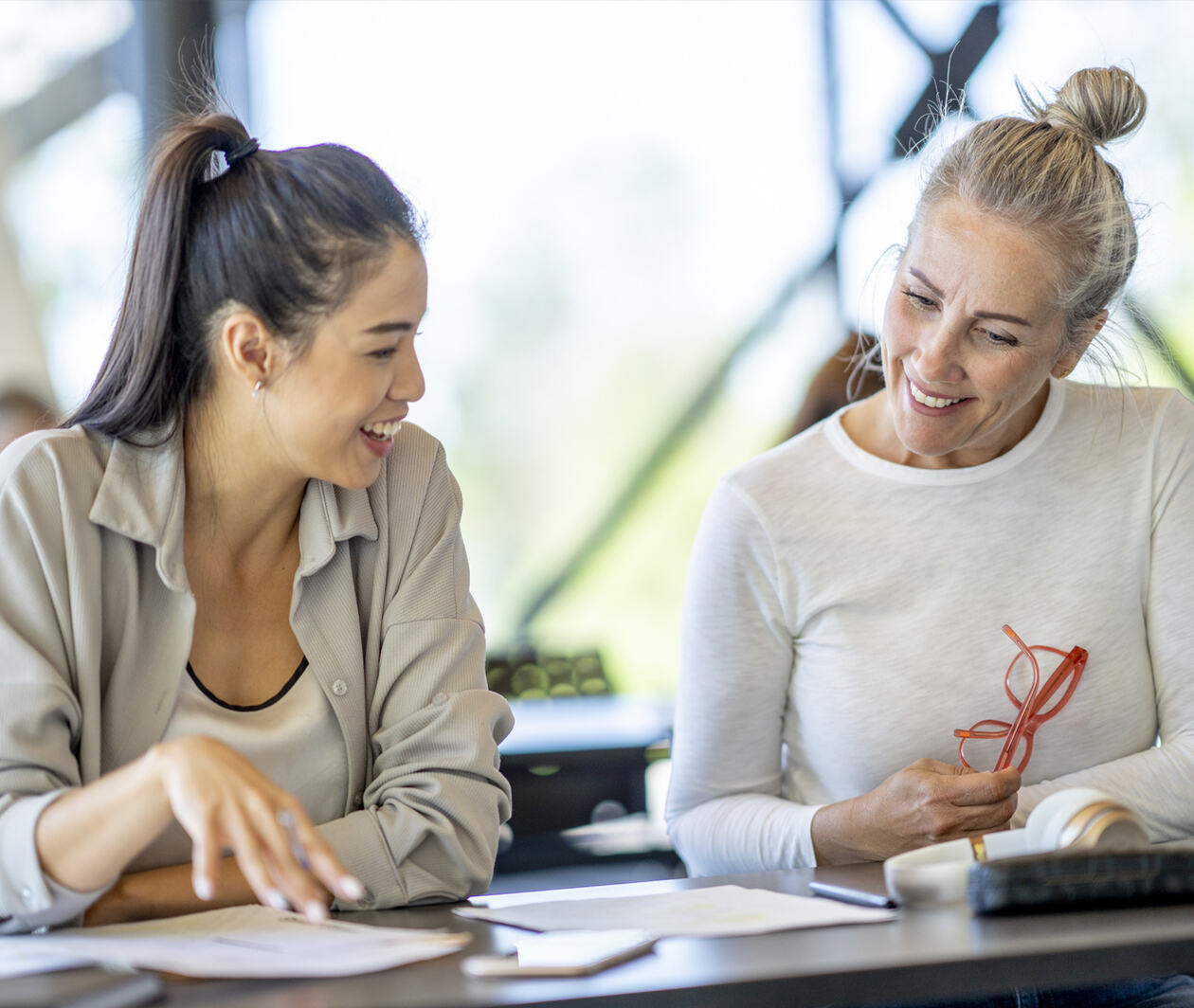 An asian women sit at a desk together with a caucasian woman as they meet to discuss various papers and reference materials out in front of them. 