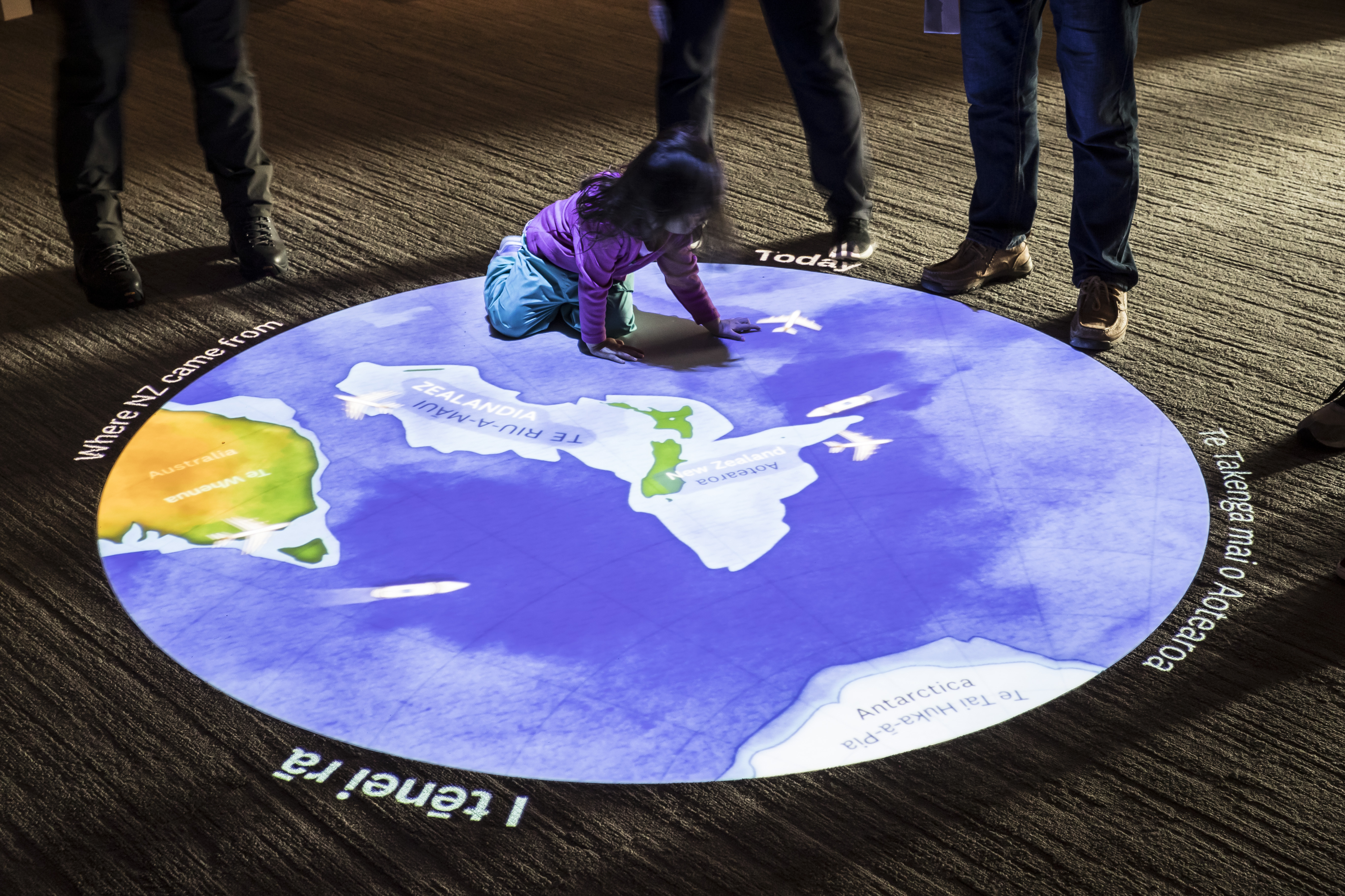 A young girl with dark hair and a purple jacket kneels on the floor engaging with a circular projection onto the ground beneath her showing New Zealand from above with the larger white continent of Zealandia illustrated under the sea.