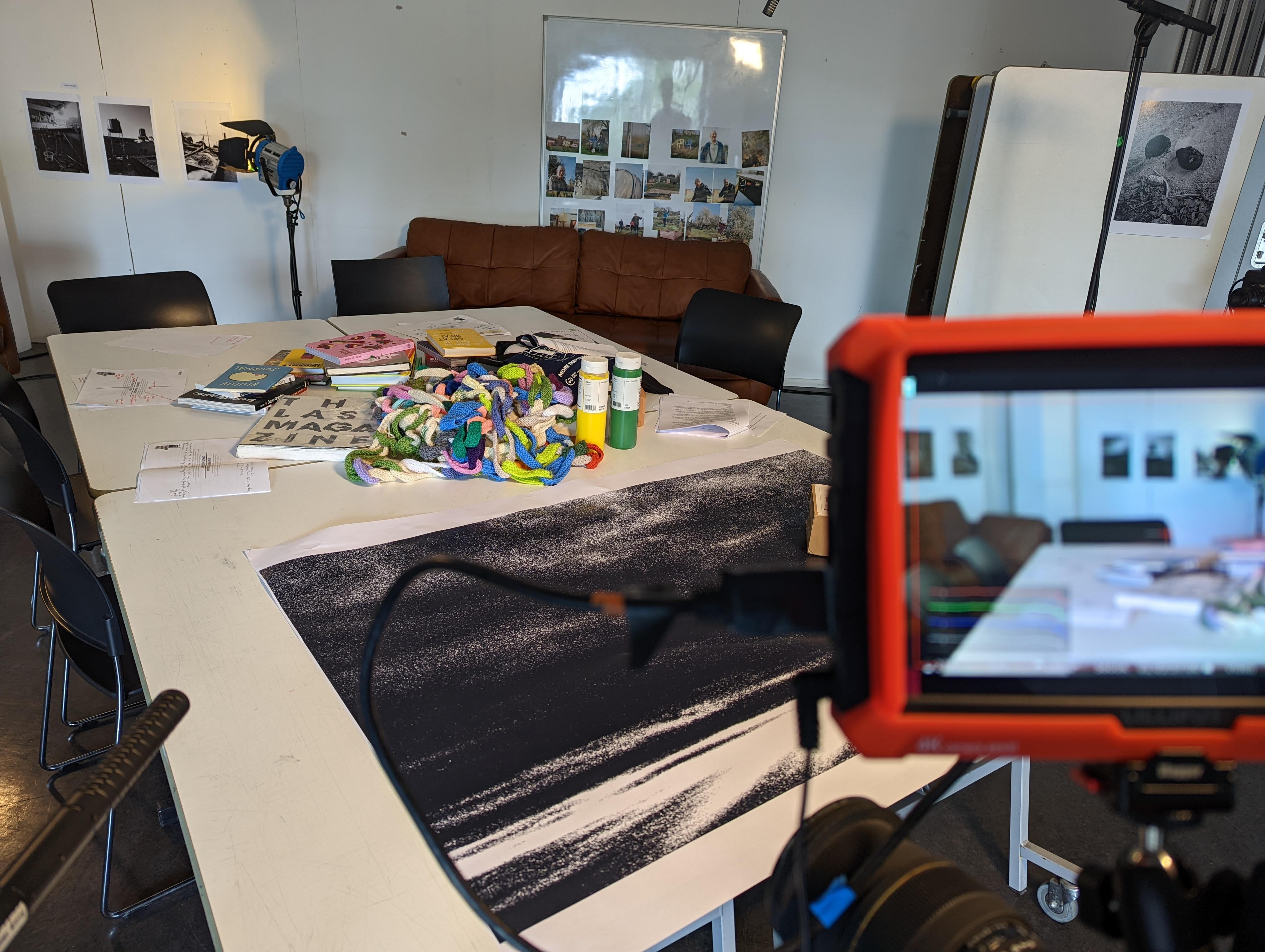 Large white table covered in books and a multicoloured crochet chain and a big black and white print, there is a red camera screen in the foreground.