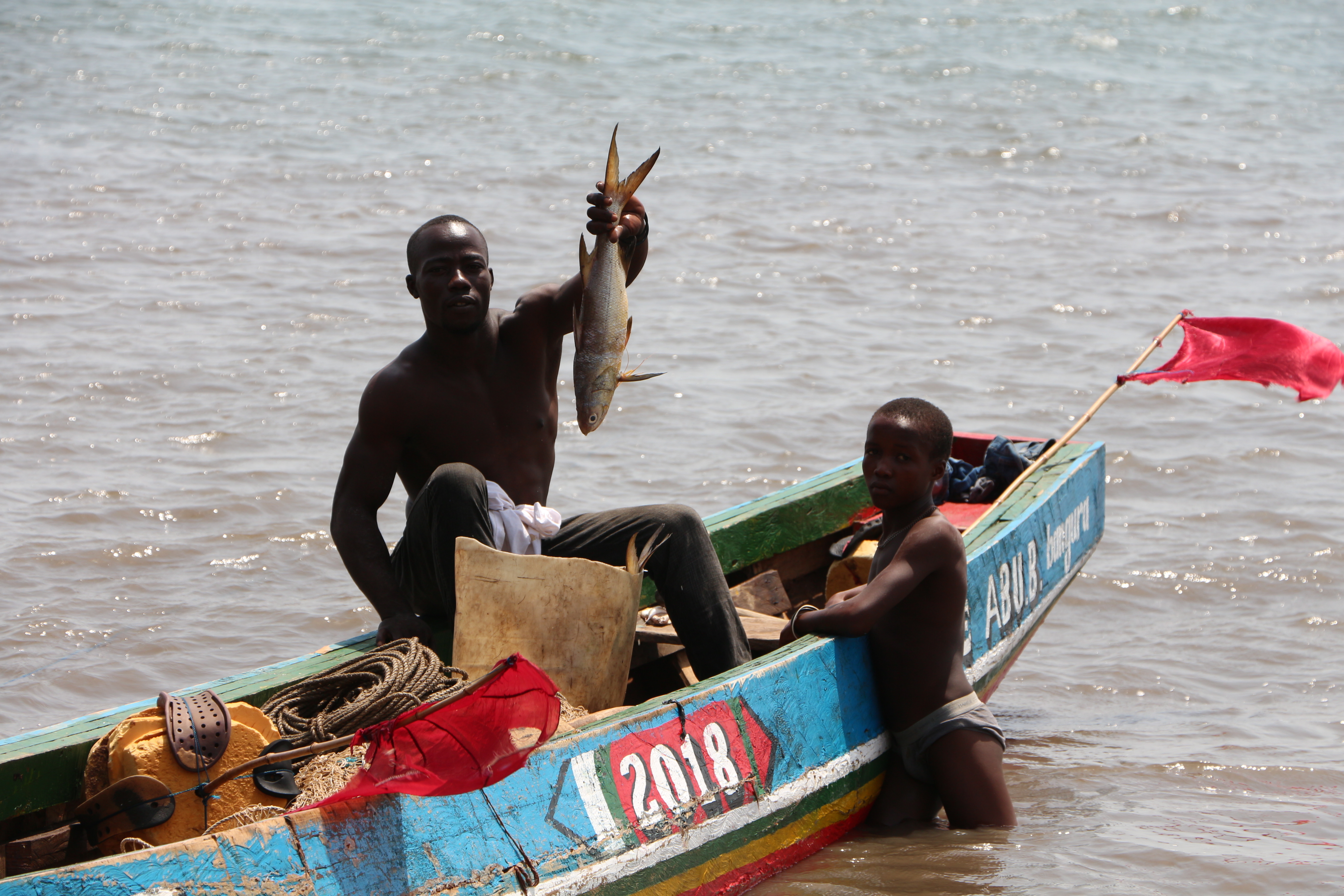 A man and youth on a blue boat, sitting in the sea. The man is holding up a fresh fish.