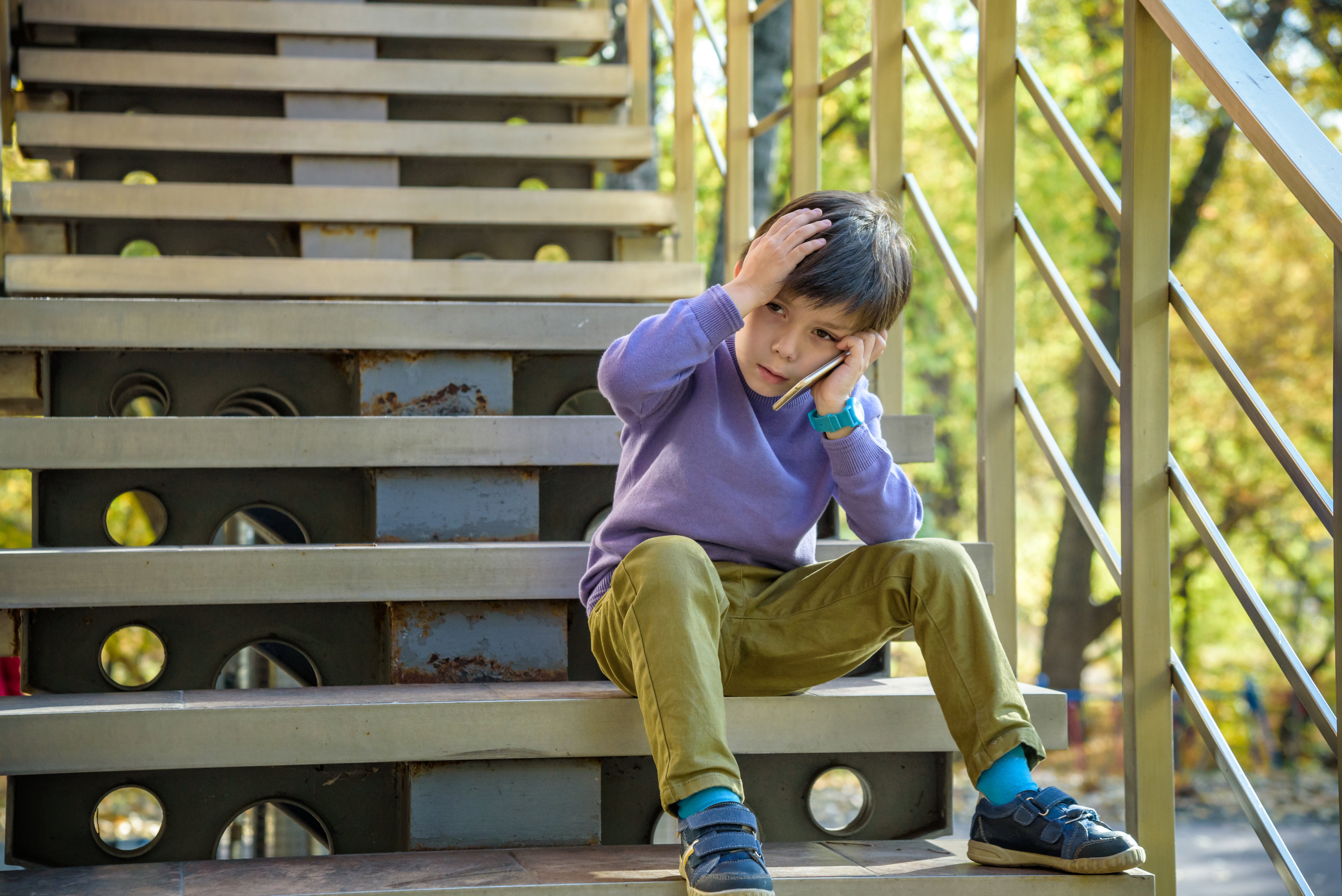 Boy sitting on outside steps with a hand to his head and the other hand holding a phone which he is looking at