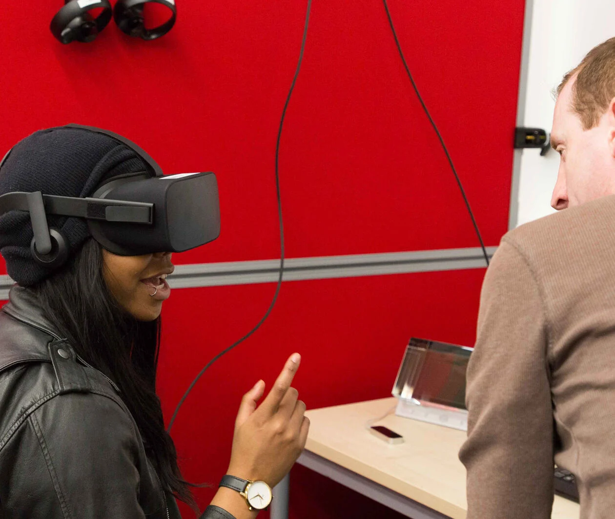 A woman in a virtual reality headset sitting a desk with a man nearby talking to her