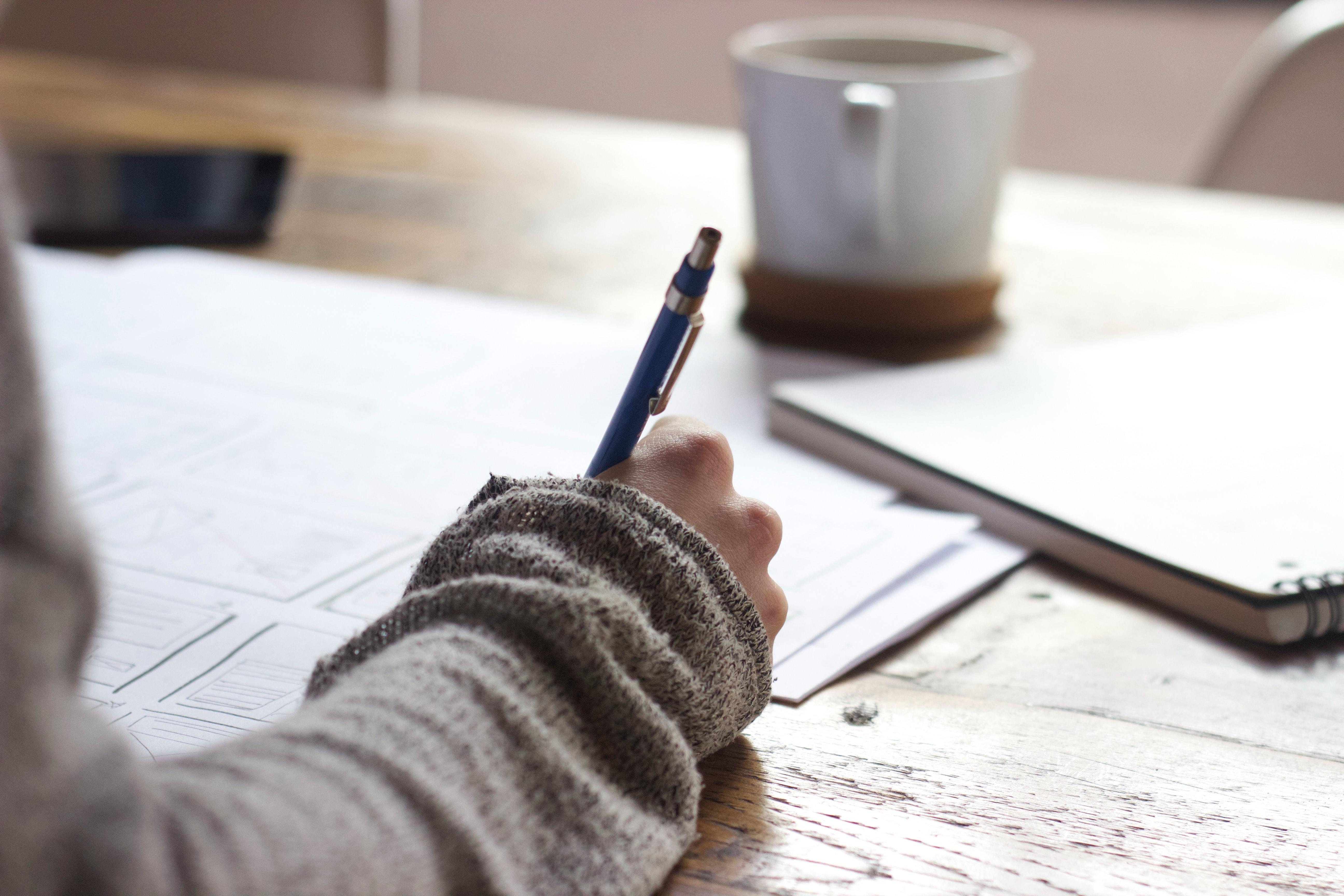 person writing on brown wooden table near ceramic mug