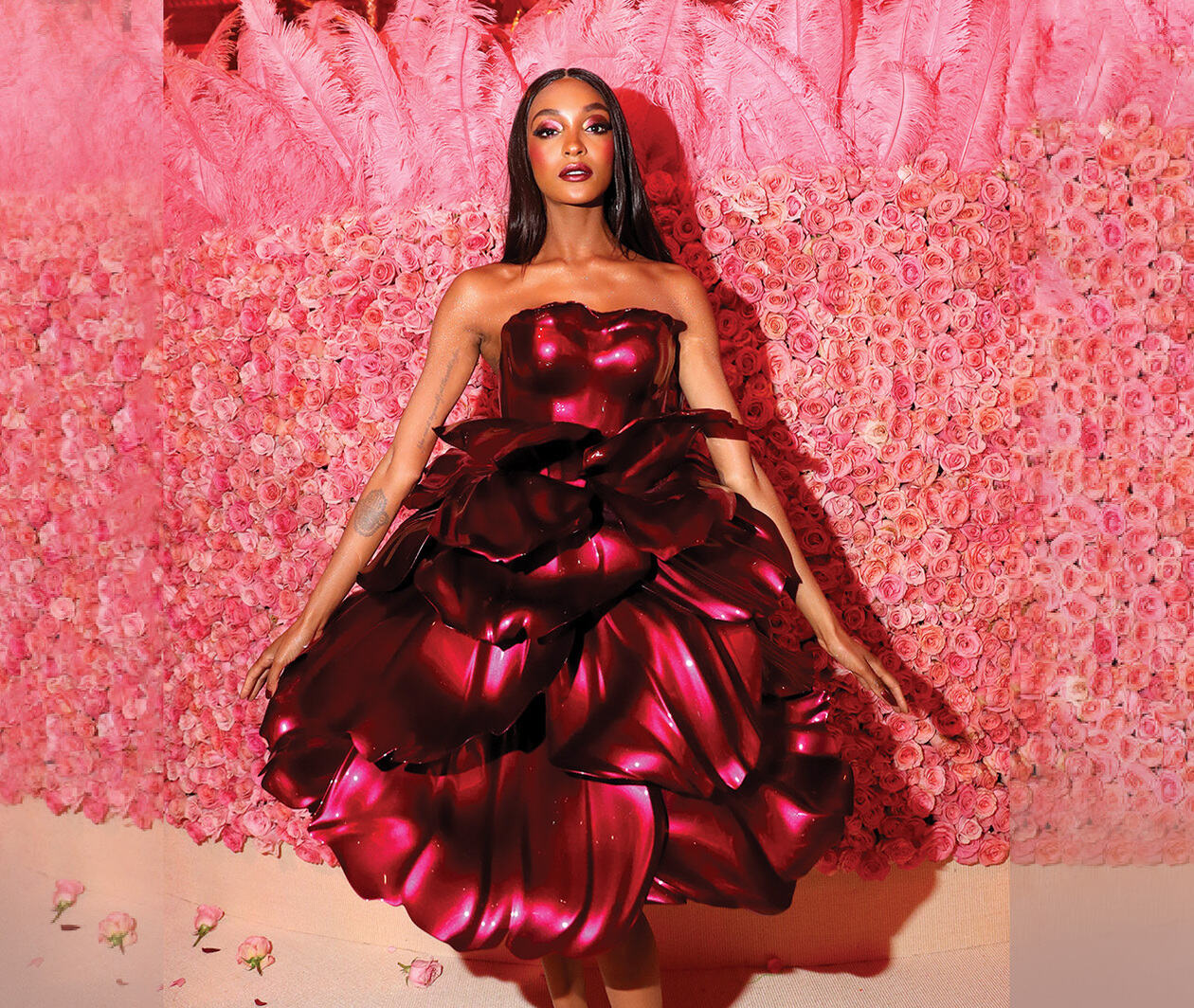 A model wearing a long red dress poses in front of a flower wall.