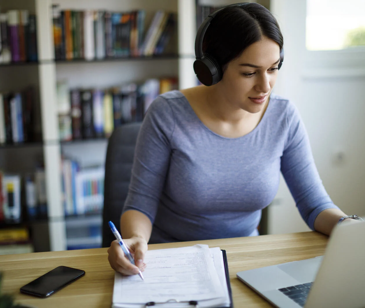 Woman sat at a laptop, while holding a pen over a notepad, wearing headphones