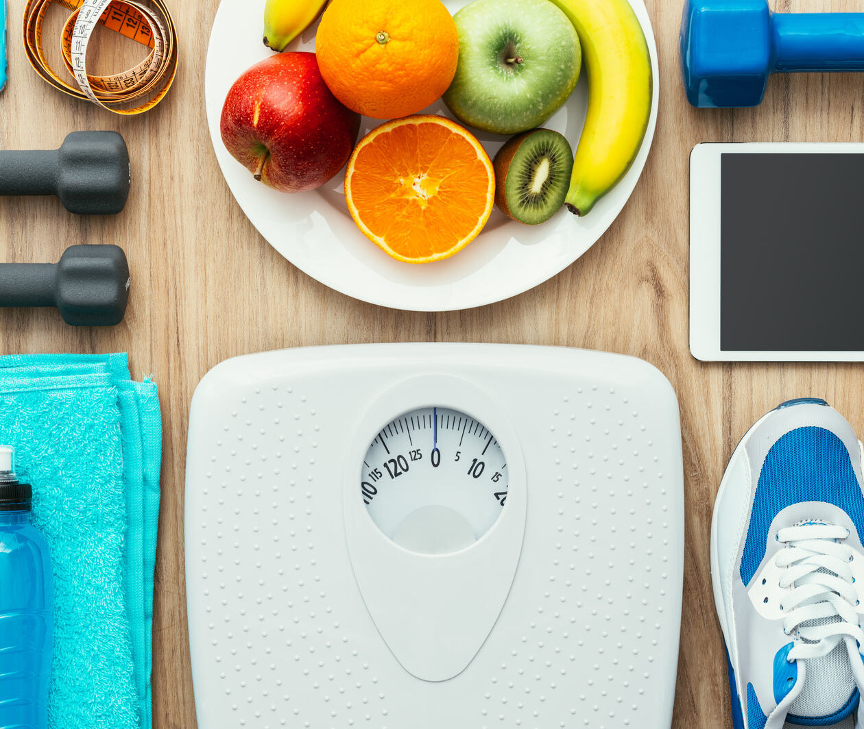 Laid on a brown wooden floor are: a set of white scales, a blue towel, a water bottle, a pair of blue and white trainers, a bowel of fruit, an iPad, an iPhone, a measuring tape and some small weights. 