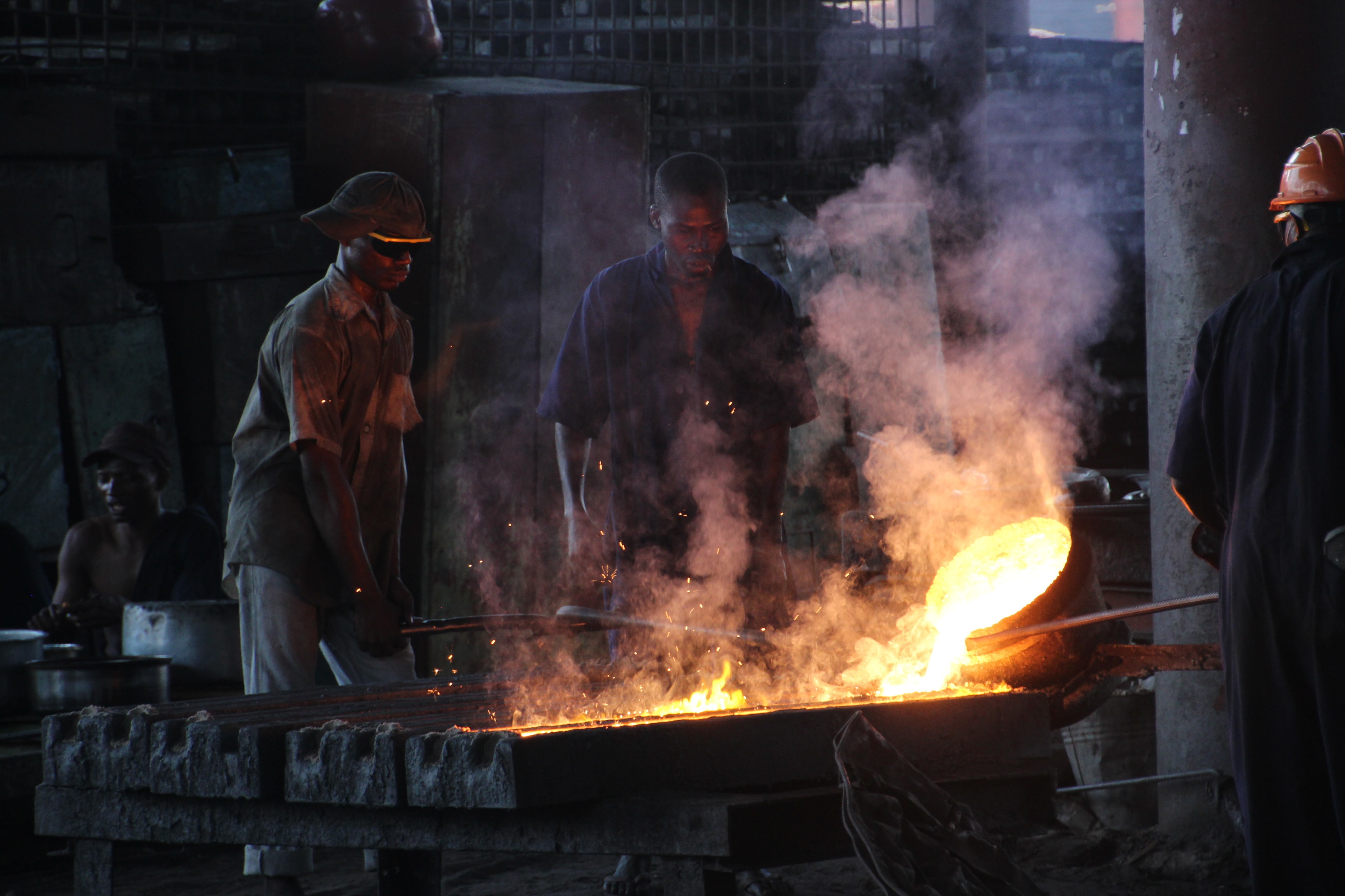 Iron foundry workers are standing around the oven, and we see they are melding metal, and fumes are in the air. The workers are very close to the oven, poking with metal bars into the melted substance. They do not have respiratory protective equipment.