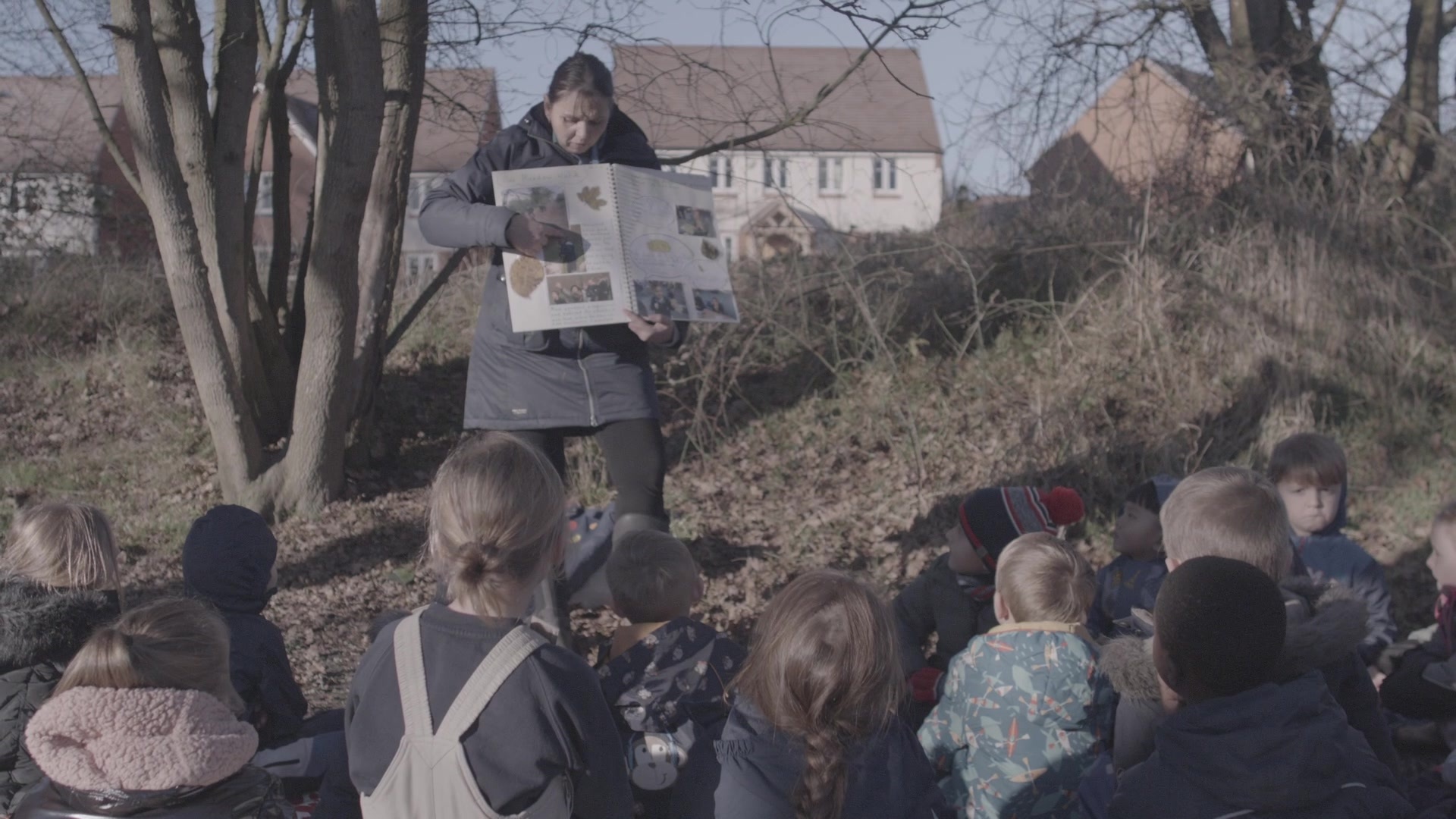 A teacher pointing to a large book to a class all sitting outside