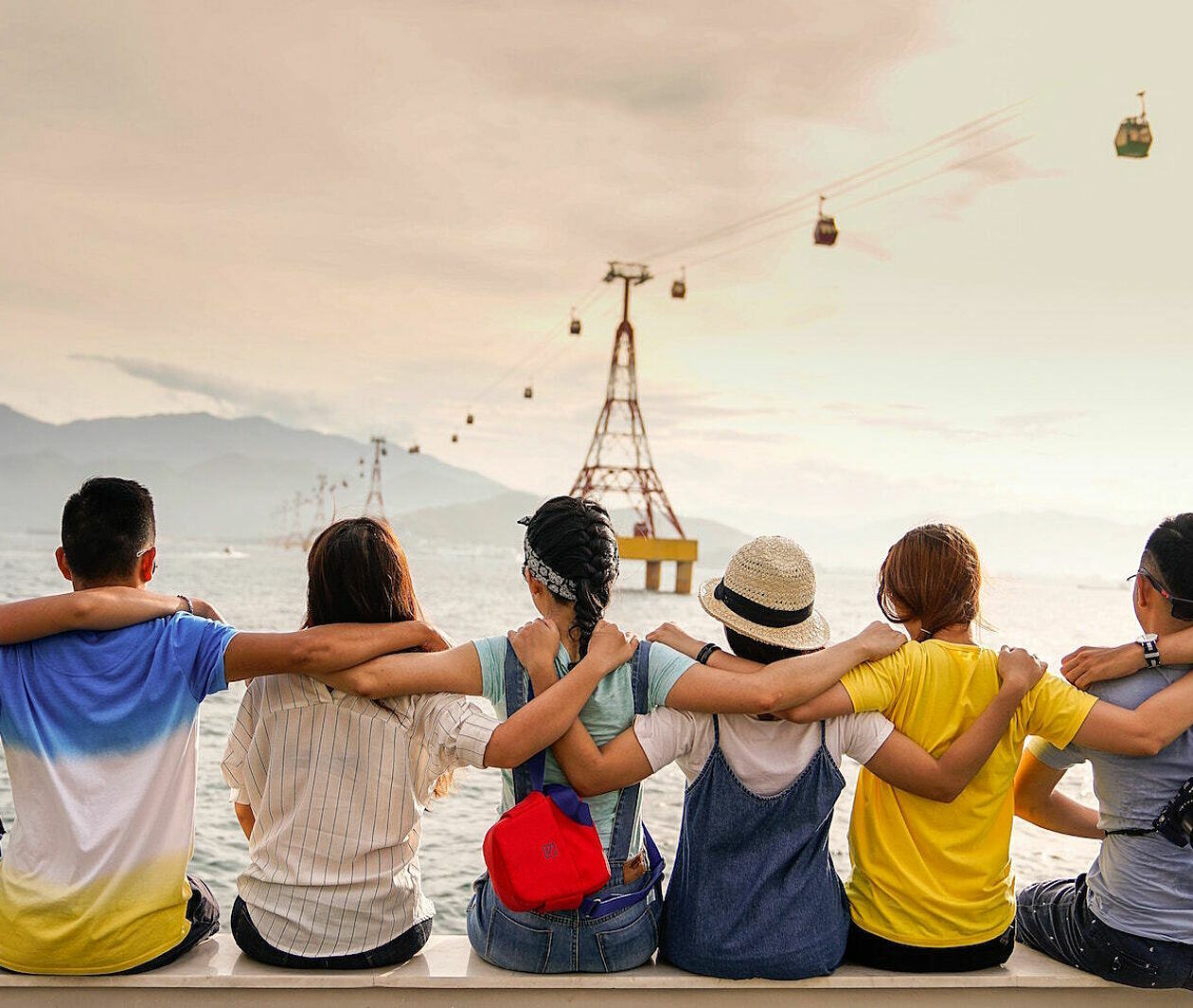 A group of people, facing away from us, siting on a quayside, looking out over a bay, with their arms interlinked