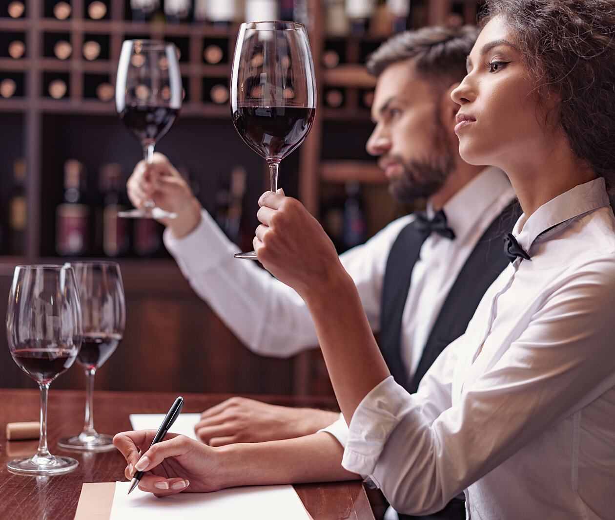 A man and woman in smart attire stand at a table holding glasses of red wine in front of them making notes on a notepad