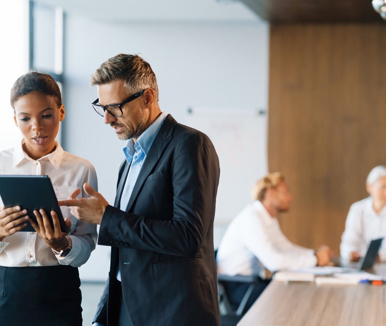 A man and woman in business attire looking at a tablet together in a conference room with people behind them