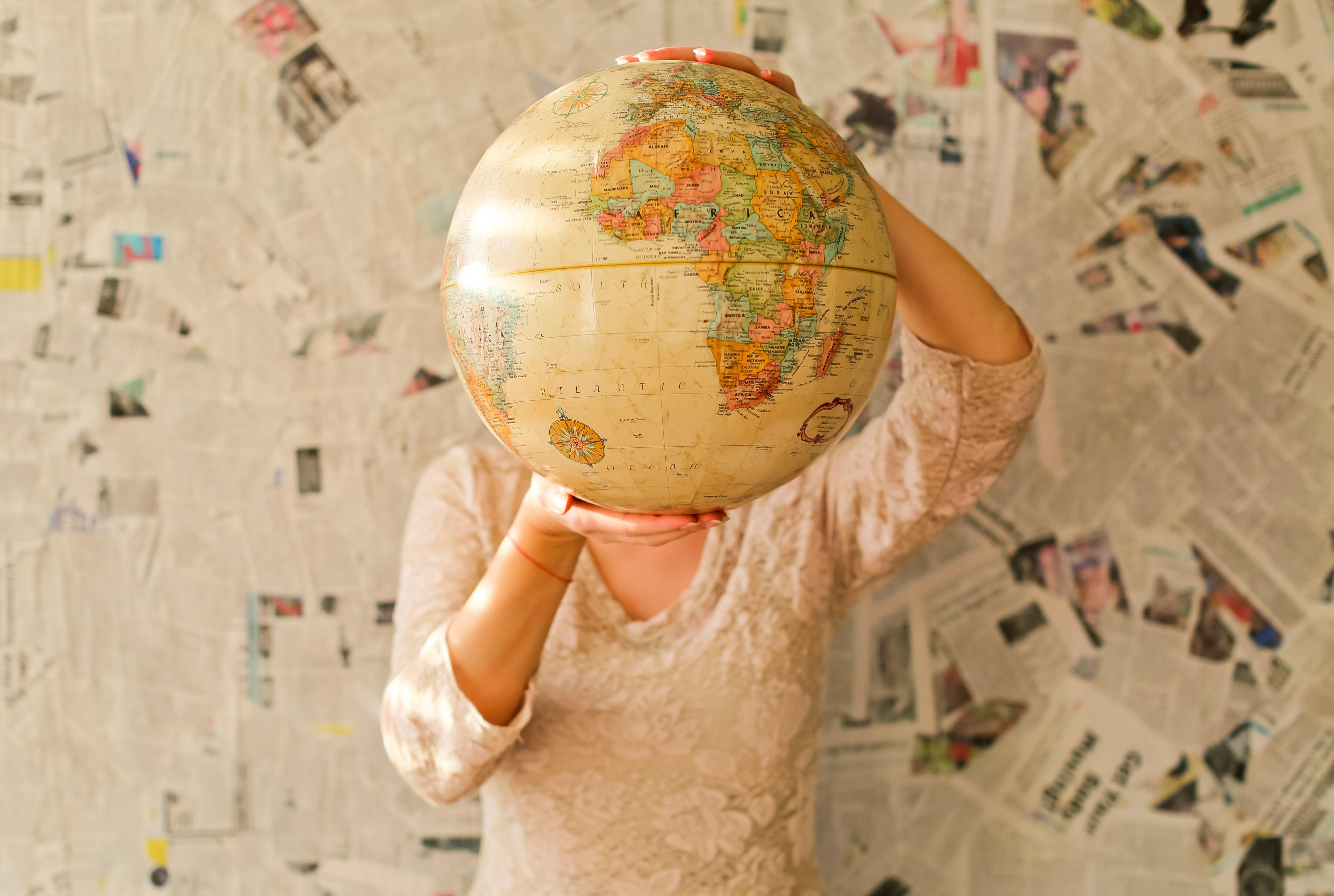 Two hands holding a large globe in front of the face of a person: torso and legs visible on a background of newspaper clippings.