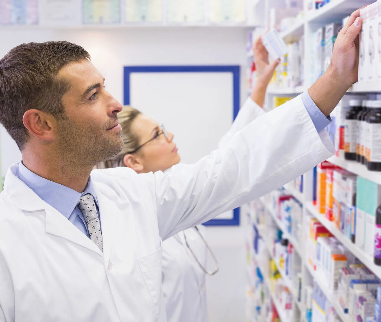 Two pharmacists reaching for medicines on a shelf.