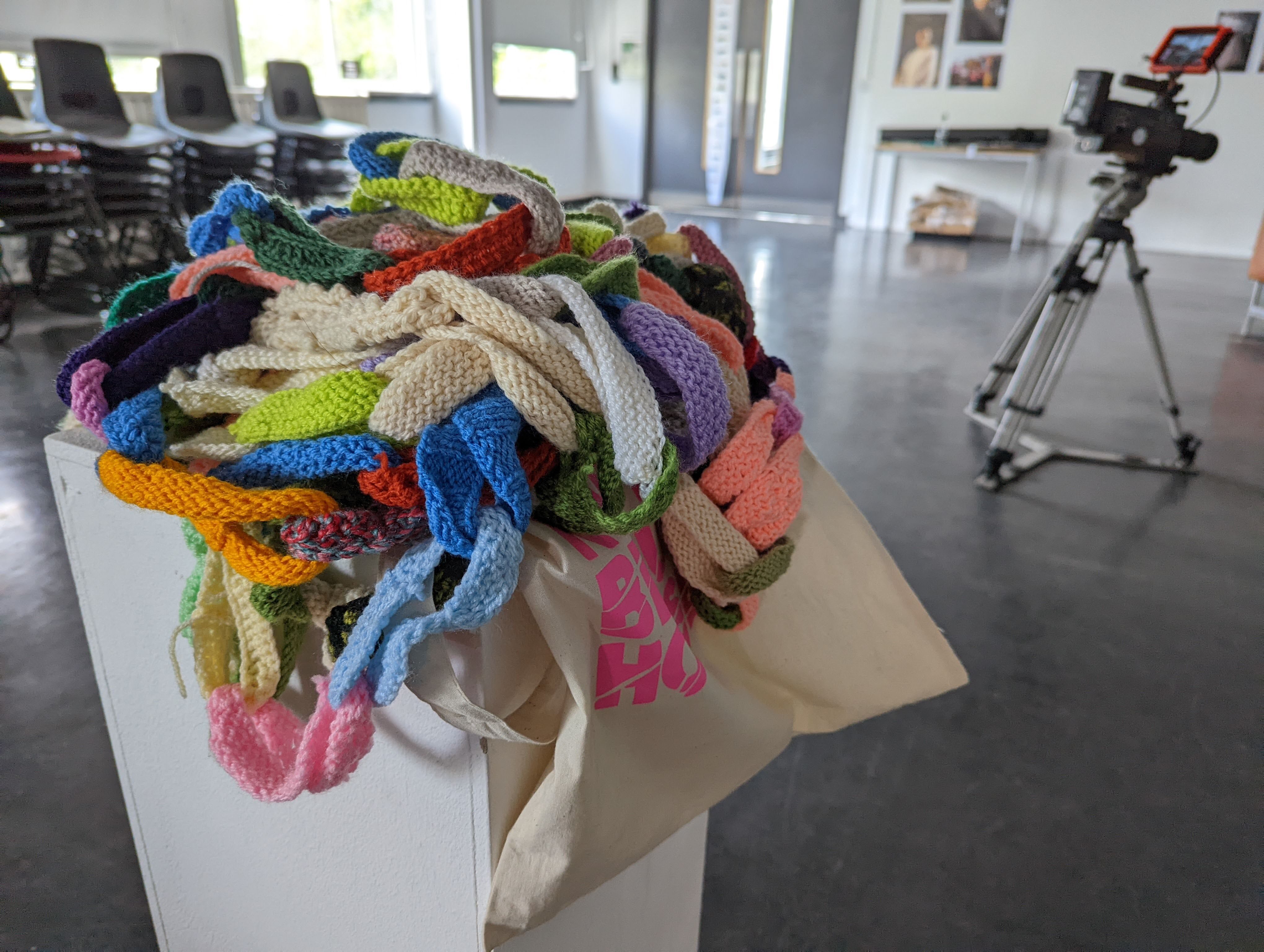 Multi coloured crochet chain set up on a white plinth, there is a camera and some stacked chairs in the background.