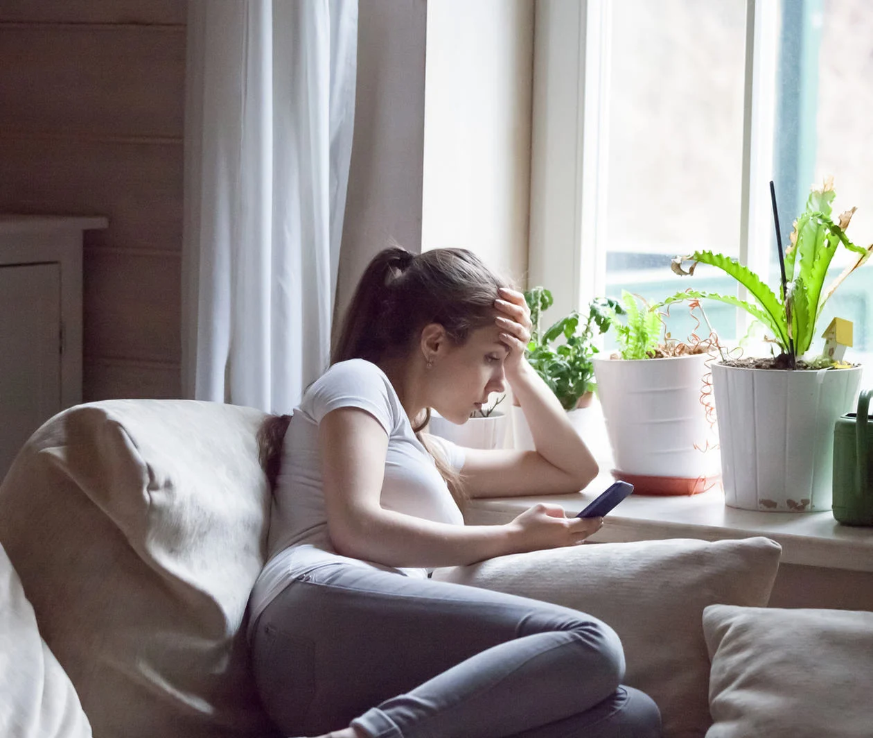 A teenage girl sits by the window, looking at her mobile phone, head in her hand