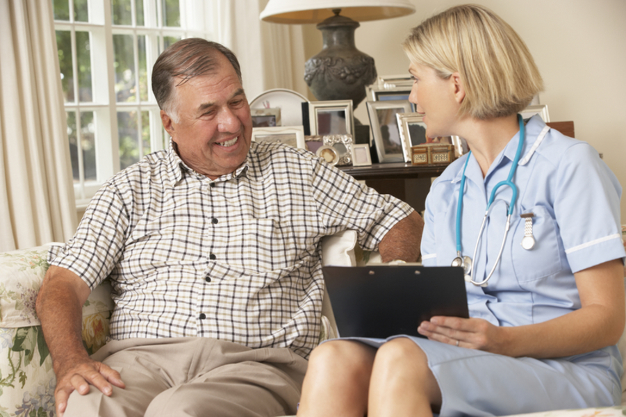 A nurse sitting talking to a man in a sitting room with a clipboard in her hand.