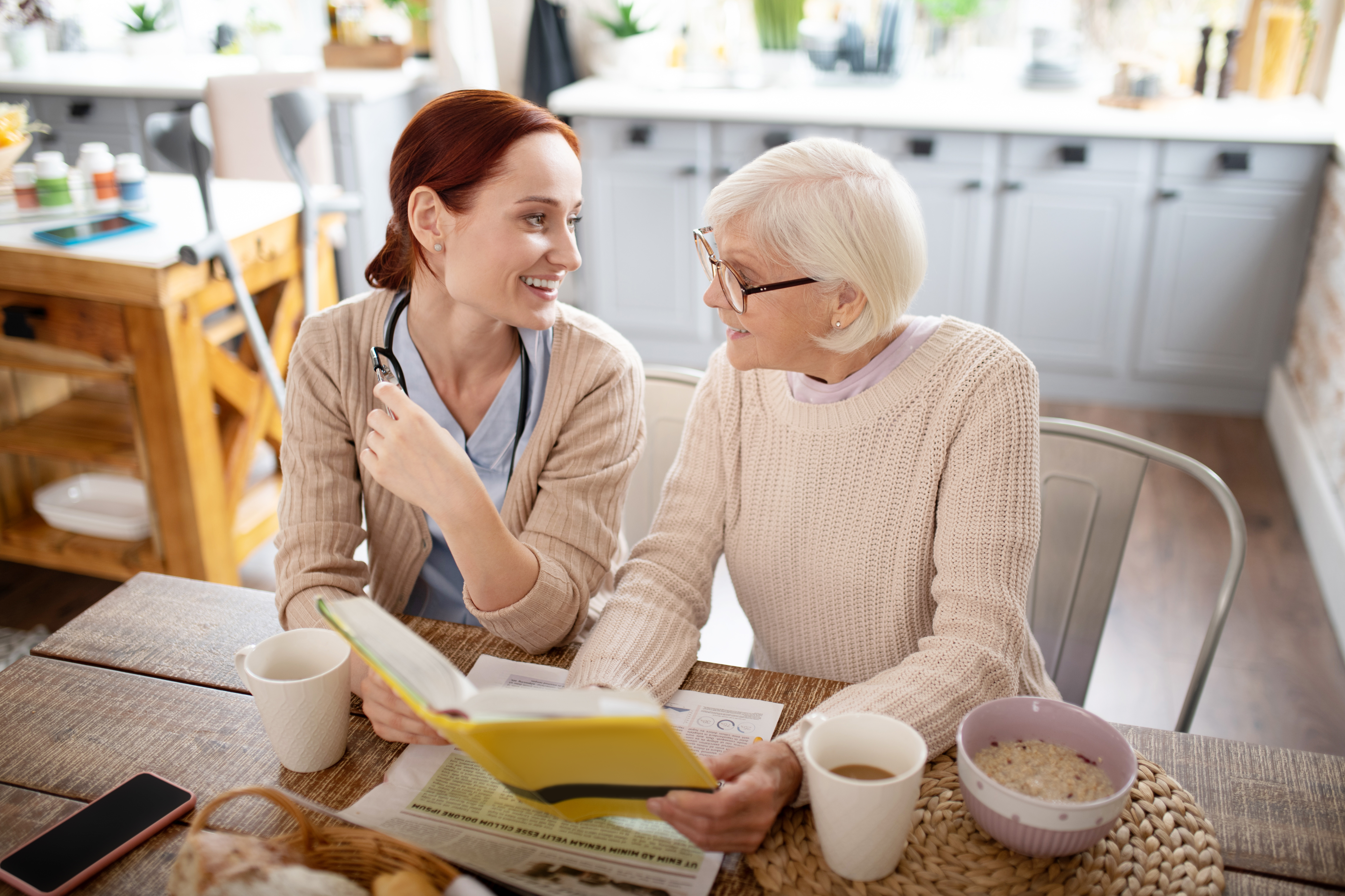 A young healthcare worker sits and talks with mature woman looking at a book in a kitchen.