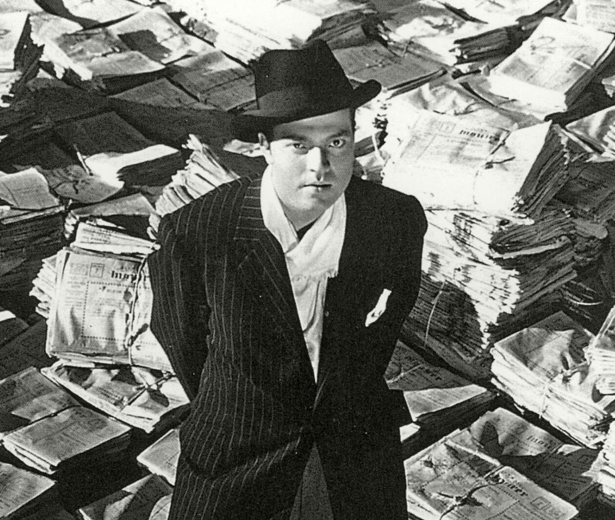 man stands on a mound of newspapers looking directly at the audience. A still image from Citizen Kane.