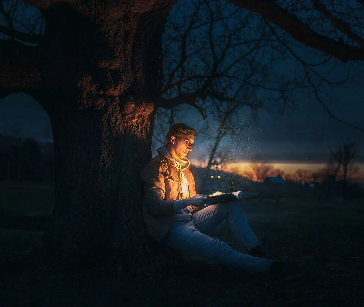 Young person with glasses sitting at the base of a tree with an open book that is illuminating their face