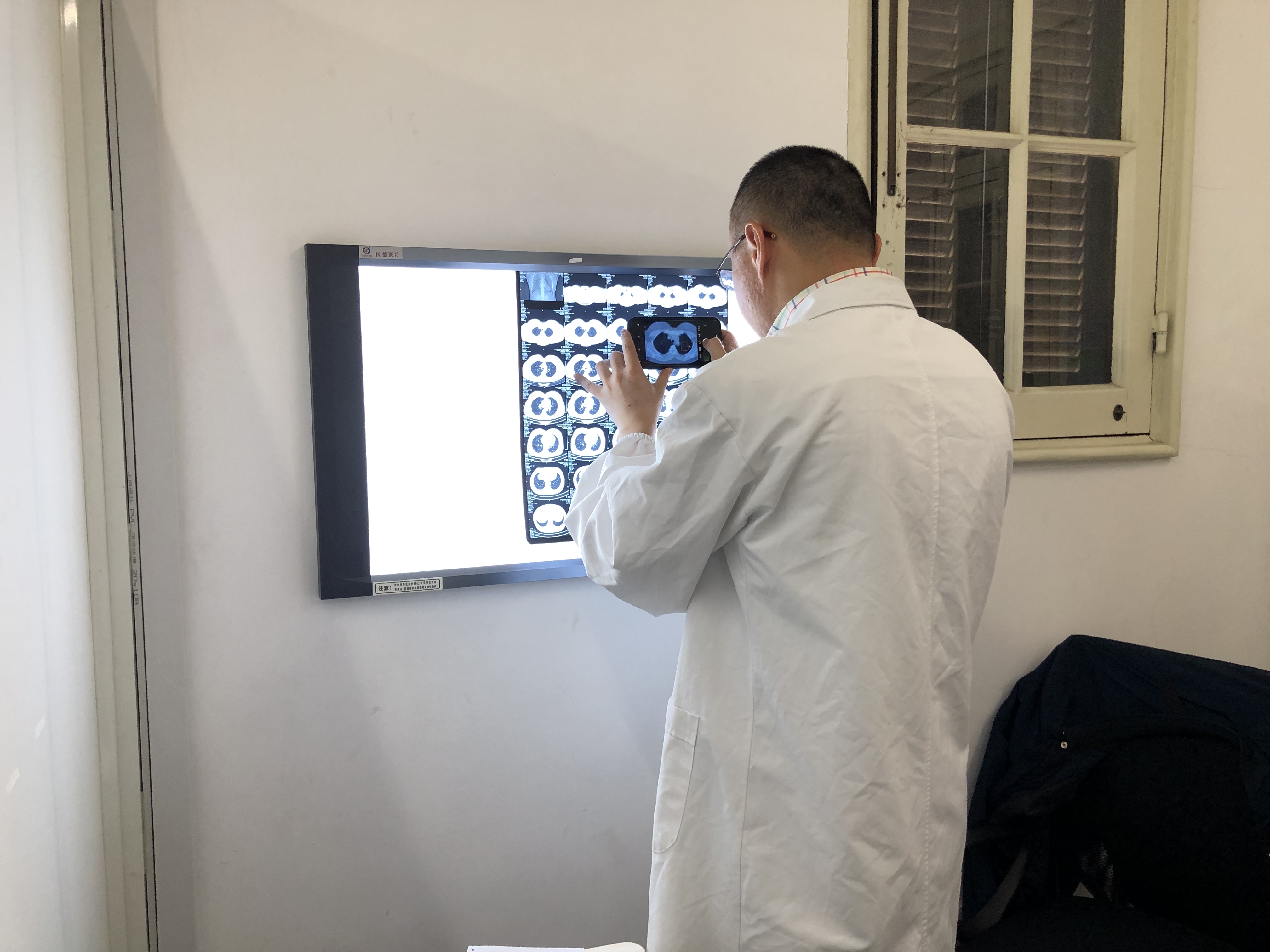 A Chinese doctor with his back to the camera holds his smartphone up to take a photo of some CT scans of lungs which are pinned to a medical illuminator type device. A window can be seen to the right.