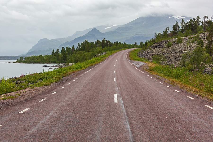 A road leading past a lake and into a forest with mountains in the background.