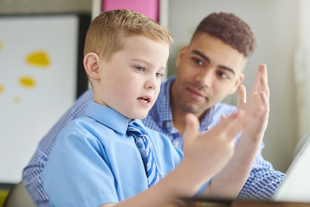 A male teacher sitting with a young male pupil. The teacher is holding a pad, and the pupil is looking at his hands.