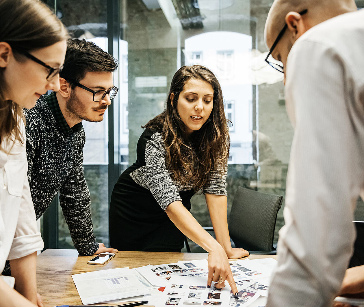 Work colleagues gathered around a table. A woman is pointing at an image on a document.