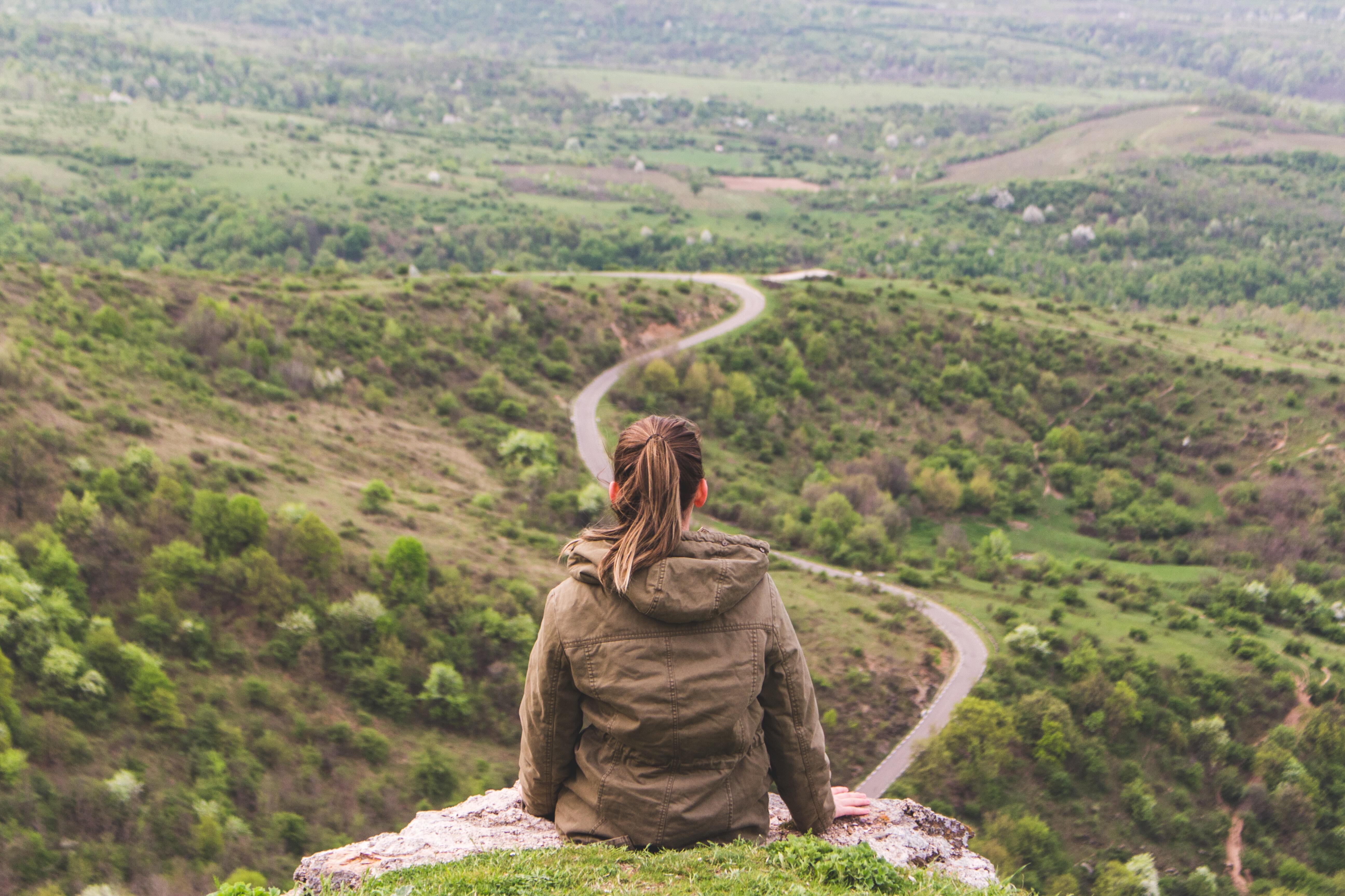 Woman sat on grey cliff looking out over landscape with a winding road