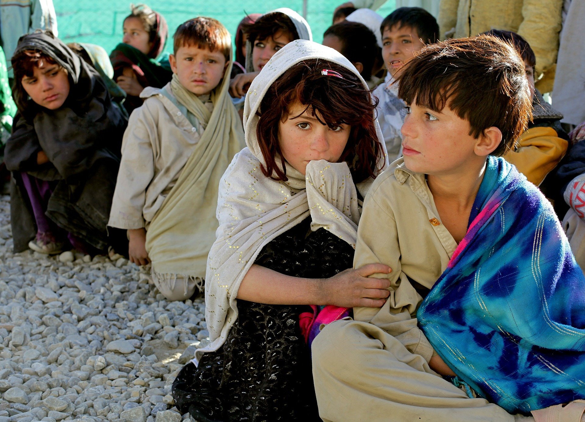 A group of children sitting wrapped with shawls on pebbles.