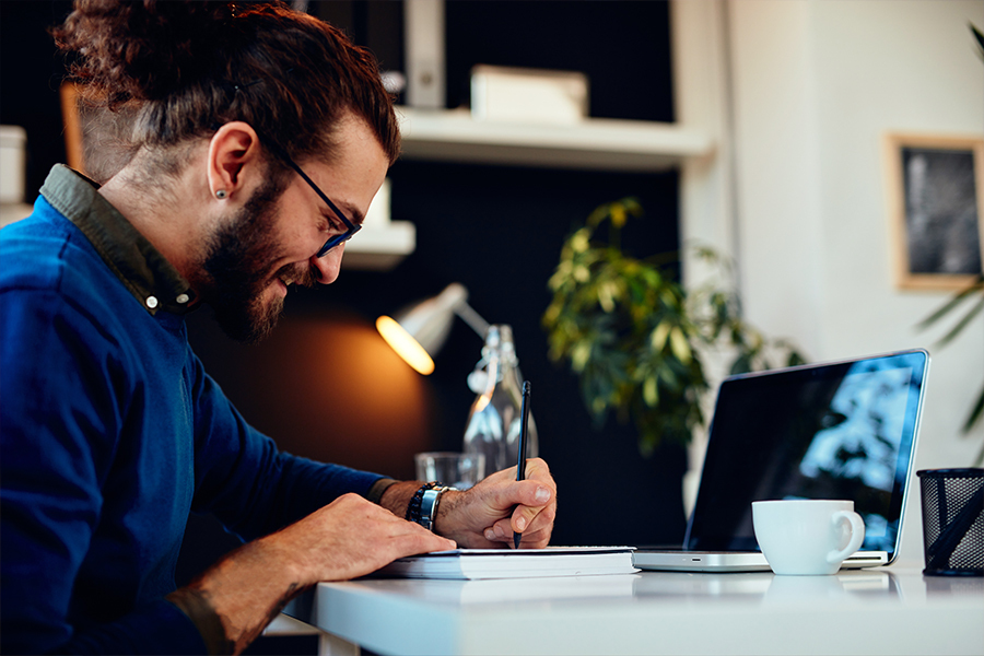 A smiling man sitting by a table taking notes in his notebook.