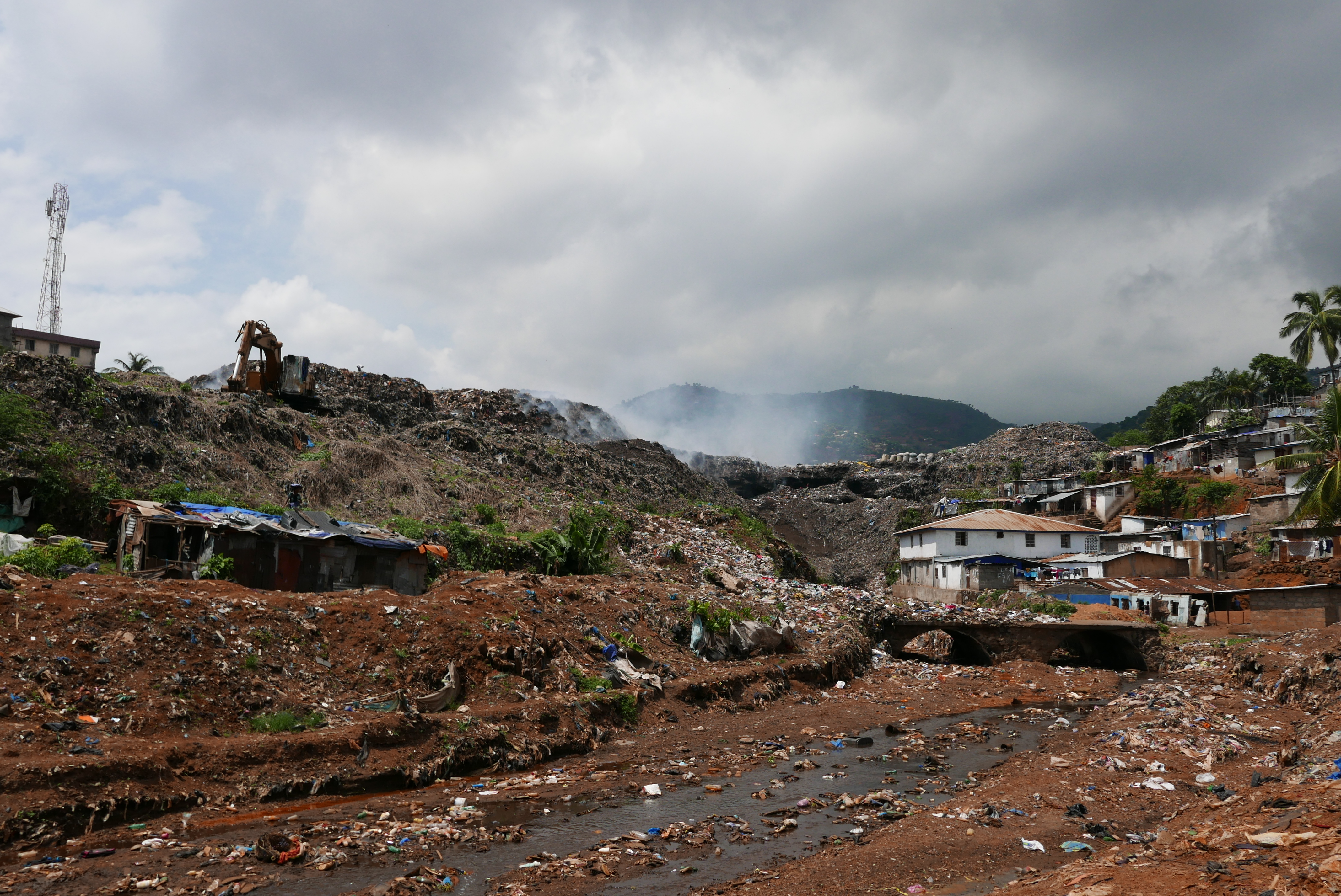 Image shows a large rubbish heap with houses on one side and smoke rising above it.