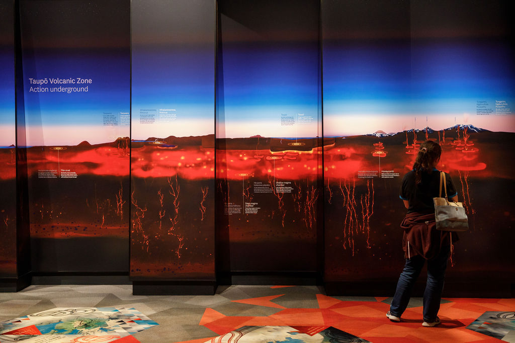 A woma, dressed in dark clothing stands with her back to us looking at 4 large wall panels, appearing to lean on alternating angles, depicting a cross section of magma activity beneath the ground in Taupō, against a blue sky.